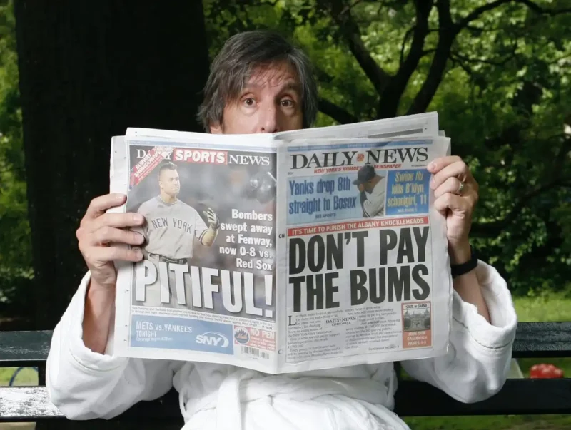 Andy Borowitz seated on a park bench wearing a white bathrobe. He's looking surprised peeking over a newpaper, with headlines "Don't pay the bums" and "pitiful!"